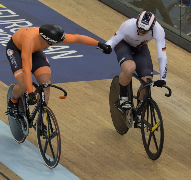 Zwei Männer mit Helmen fahren Fahrräder auf einer Velodrombahn mit einer Glaswand im Hintergrund.