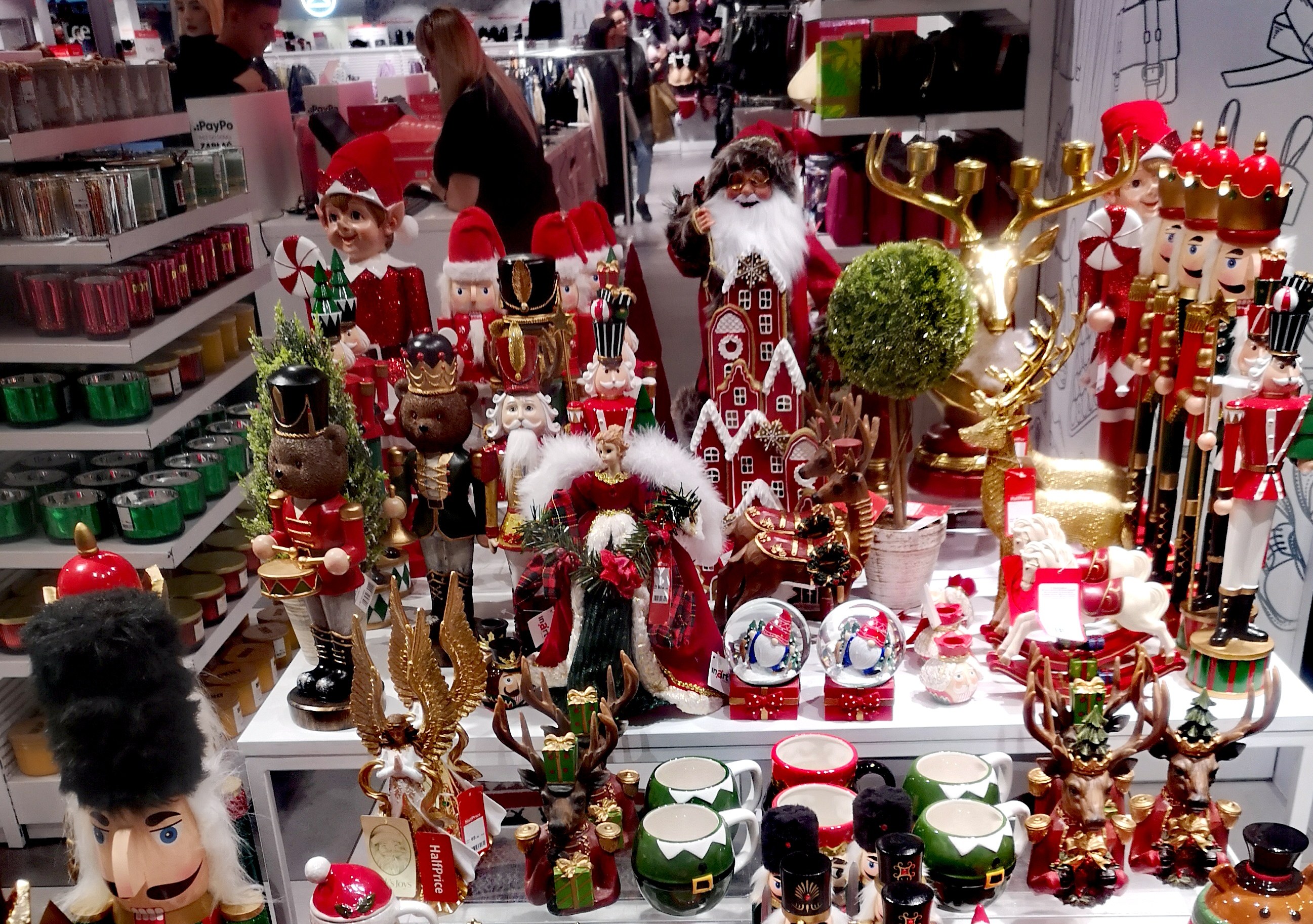 Weihnachtsdekorationen-Ausstellung mit Spielzeug, Tassen und anderen Gegenständen auf Tischen und Regalen, mit Menschen und Lichtern im Hintergrund.