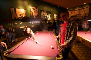 In einem Club spielen zwei Personen Snooker an einem von zwei Tischen, während mehrere andere im Hintergrund stehen und reden, vor einer cremefarbenen Wand mit Fotopostern.