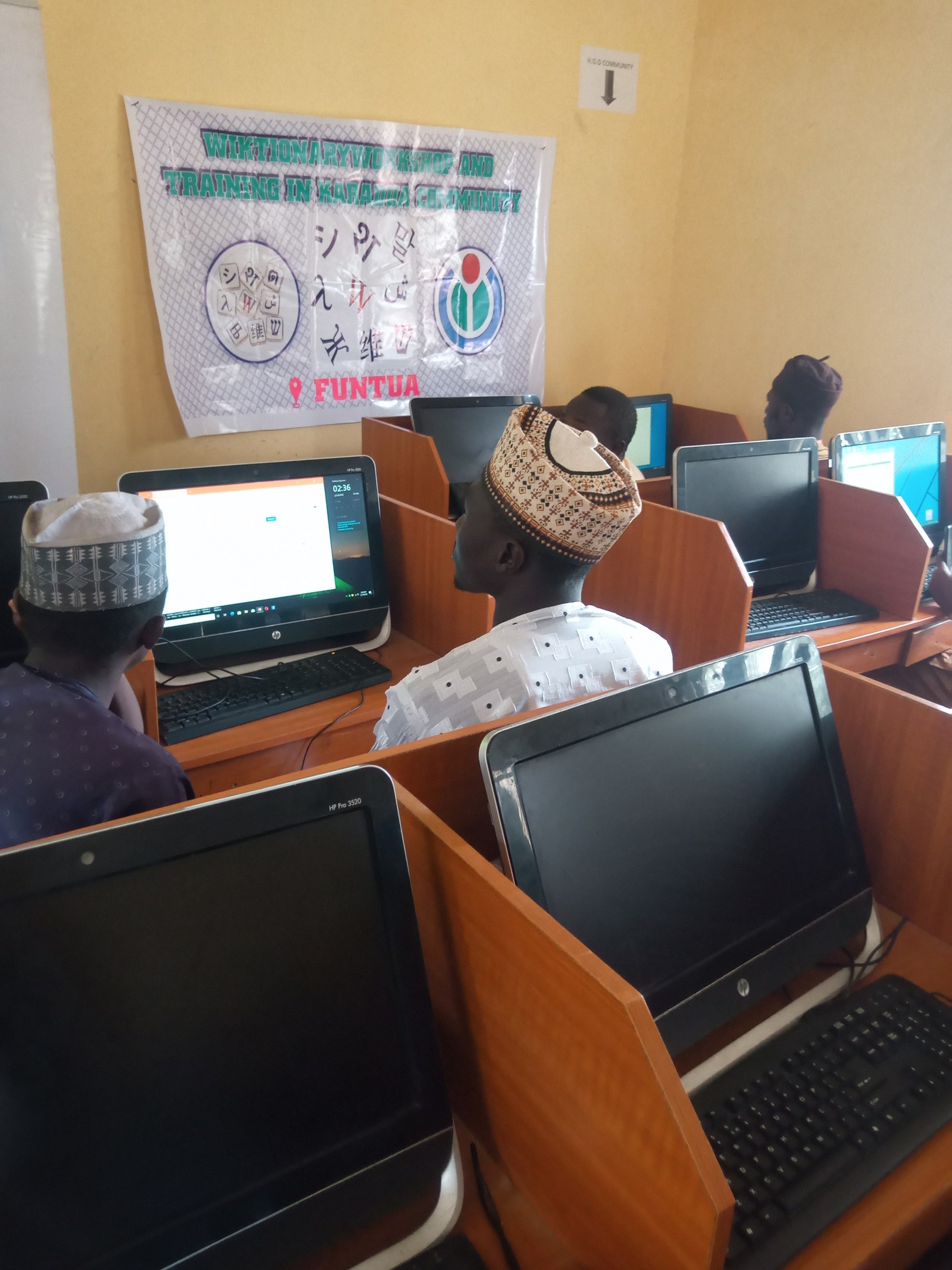Gruppe von Menschen an Tischen mit Computerbildschirmen, Tastaturen und Mäusen bei einem Workshop über Computertechnologie, mit einem Plakat im Hintergrund.