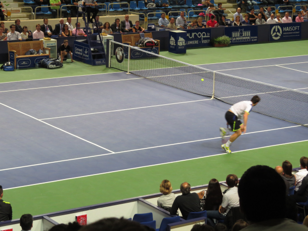 Ein Mann spielt Tennis auf einem Platz mit einem Ball in der Luft, der einen Schläger hält, vor einem sitzenden Publikum, mit einem Netz, Bannern und Treppen im Hintergrund.