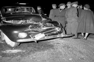 Ein Schwarz-Weiß-Bild eines beschädigten Autos am Straßenrand, umgeben von einer Gruppe besorgter Menschen.