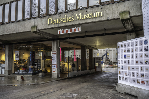 Außensicht des Deutschen Museums in Berlin, das einen großen Bau mit Glasfenstern, Säulen und einer Namenschilder zeigt, neben einer Informationsanzeigetafel, mit anderen Gebäuden, Bäumen und Himmel im Hintergrund.