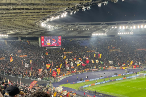 Eine große Menschenmenge in einem Stadion mit schwingenden Flaggen, Gras, einem Netz und Schildern mit Text auf dem Boden, viele Zuschauer in den Rängen, einige mit Mützen, ein Bildschirm im Hintergrund und beleuchtete Deckenlampen, die eine Atmosphäre der Aufregung und Vorfreude auf ein Fußballspiel schaffen.