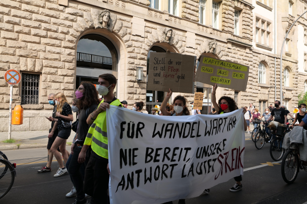 Eine Gruppe von Menschen marschiert auf einer Straße, einige halten Protestschilder und andere fahren Fahrräder, mit einem historischen Gebäude mit gewölbten Fenstern, Säulen und Skulpturen im Hintergrund.