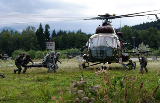 Soldaten tragen einen Mann auf einer Trage vor einem Hubschrauber, mit Gras, Pflanzen, Bäumen, Gebäuden, Hügeln und einem bewölkten Himmel im Hintergrund.