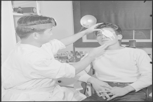 Ein Mann sitzt in einem Stuhl, während ein Arzt ihn mit einem tragbaren Instrument untersucht; im Hintergrund ist ein Fenster mit einem Vorhang und einige Gegenstände zu sehen.