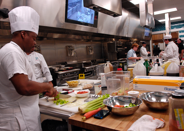 Eine Gruppe von Köchen in einer Küche, die Essen auf einem Schneidbrett zubereiten, mit dem Koch in der foreground, der eine weiße Schürze und eine Mütze trägt und ein Messer hält, umgeben von verschiedenen Utensilien, Schüsseln, Gemüse, einem Mobiltelefon, Flaschen und anderen Gegenständen, mit einem Fernseher und Deckenleuchten im Hintergrund.