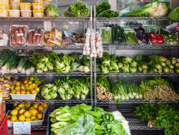 Ein Supermarktwarenregal mit frischem Gemüese wie Gemüese und Obst in Plastikverpackungen, zusammen mit Kisten und einer Schautafel und einem Glasfenster im Hintergrund.