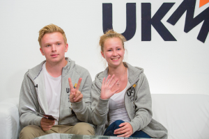 Ein Paar in passenden Jacken auf einem Sofa sitzend mit 'UKM' im Hintergrund.