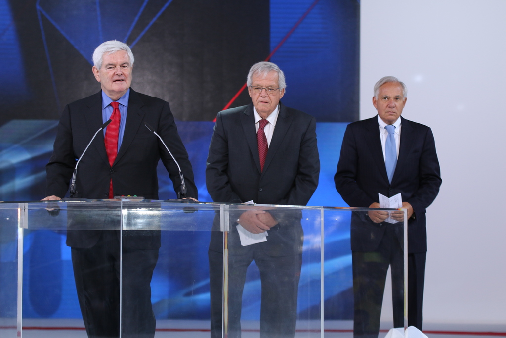 Ein älterer Mann in einem schwarzen Mantel und roter Krawatte steht an einem durchsichtigen Rednerpult auf einer Bühne, mit einer blauen und weißen Fahne im Hintergrund.