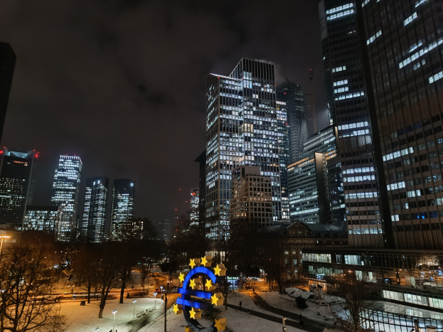 Europäische Zentralbank-Hauptquartier in Frankfurt, Deutschland bei Nacht, mit beleuchteten Gebäuden, Bäumen, Straßenlaternen, Straßenlaternen und Menschen, die auf der Straße unter einem bewölkten Himmel spazieren gehen.