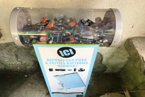 Ein Müllcontainer voller Batterien auf einem Bürgersteig, mit einem Plakat mit Text an der Wand dahinter und einer Treppe auf der rechten Seite.