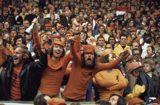 Eine große Menge in orangen T-Shirts und Hüten, die in einem Stadion jubeln, einige halten Fahnen und eine Person hält einen Megafon, mit Geländern im Hintergrund.