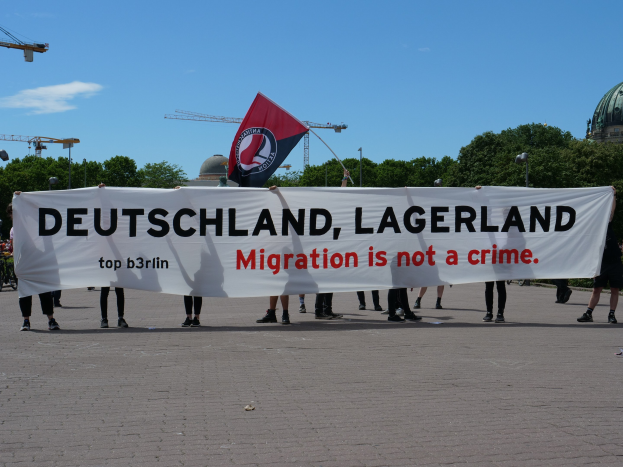 Eine Gruppe von Menschen mit einem Transparent 'Deutschland, Lagerland Migration ist kein Verbrechen' und einer Flagge, im Hintergrund Straßenmasten, Straßenlaternen, Bäume, Kräne, ein Gebäude und ein bewölktes Himmel.