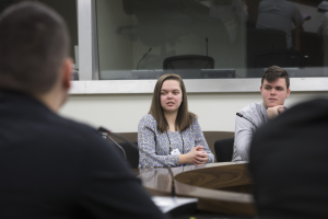 Eine Gruppe von Menschen sitzt um einen Tisch mit Papieren und einem Mikrofon vor einem Glasfenster und diskutiert.