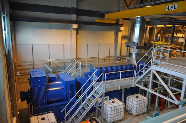 Fabrikinneneinrichtung mit Trägern, einer Treppe mit Geländer, Containern, Deckenleuchten, freiliegenden Rohren und einer Wand.