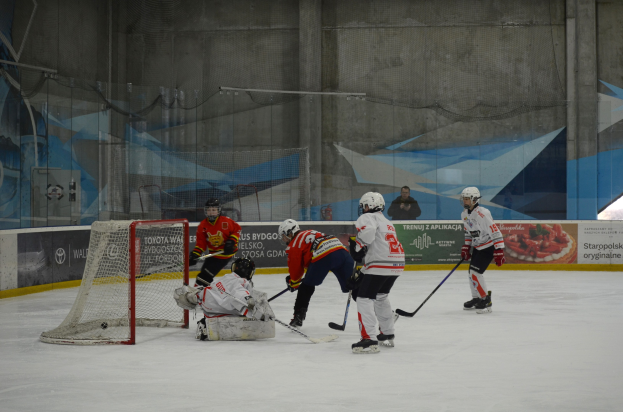 Gruppe von Menschen, die Eis hockey auf einer Indoor-Eisbahn spielen, mit Helmen und Hockey-Schlägern und einem Tor auf der linken Seite und Bannern im Hintergrund.