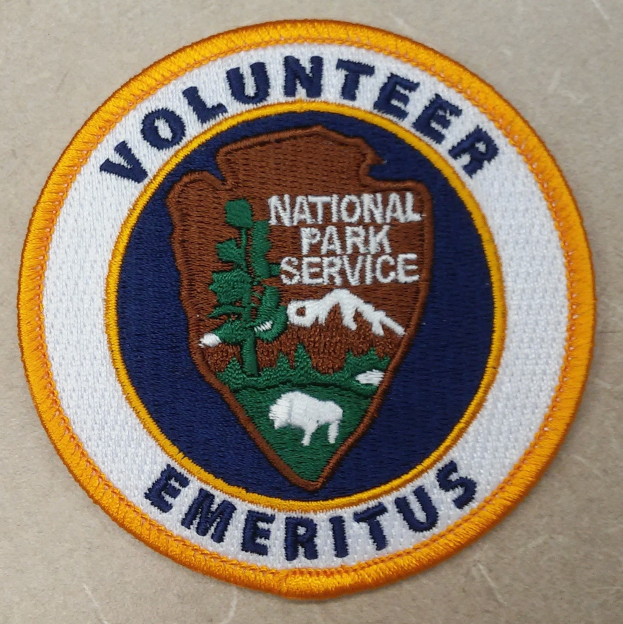 Runder Patch mit "Ehrenamtlicher Nationalparkdienst Emeritus" in fetter schwarzer Schrift auf weißem Hintergrund, umrandet von einem blauen Rand und einem gelben Kreis.