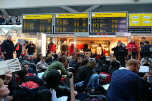 Eine große Gruppe von Menschen versammelt sich in einem Flughafen, einige sitzen mit Taschen und Papieren, andere stehen, mit Texttafeln, Schaufensterpuppen in Kleidern und Deckenleuchten im Hintergrund, was auf eine Demonstration hindeutet.