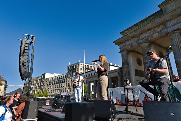 Eine Gruppe von Menschen, die auf einer Bühne Musik spielen, im Hintergrund das Brandenburger Tor, mit Instrumenten, Mikrofonen, Lautsprechern und verstreuten Gegenständen unter einem klaren blauen Himmel.
