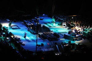 Eine Bühne mit Performern, Equipment und einer großen Zuschauermenge auf beiden Seiten in einem dunklen Hintergrund.