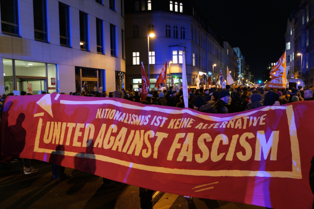 Eine Gruppe von Menschen marschiert mit einem Banner, auf dem "Einheit gegen Faschismus" steht, eine Straße entlang, mit Gebäuden, Laternenmasten und Schildern am Straßenrand und dem Himmel im Hintergrund.