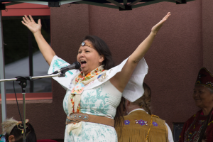 Eine Frau in einem weißen Kleid steht auf einer Bühne, schreiend in ein Mikrofon, mit einer Wand und einem Eisenrahmen dahinter.
