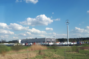 Eine Mercedes-Benz-Fabrik in Deutschland mit Fahrzeugen auf der Straße, umgeben von Gras, Pflanzen, Bäumen, Gebäuden und Polen, unter einem bewölkten Himmel.