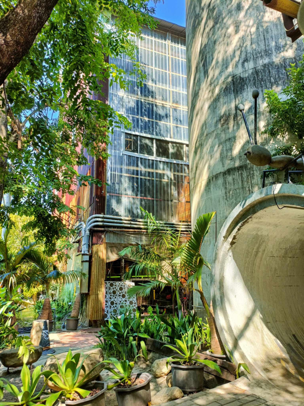 Ein Innenhof mit Topfpflanzen und Bäumen vor einem modernen, umweltfreundlichen Hotel, umgeben von üppiger Vegetation unter einem klaren blauen Himmel.