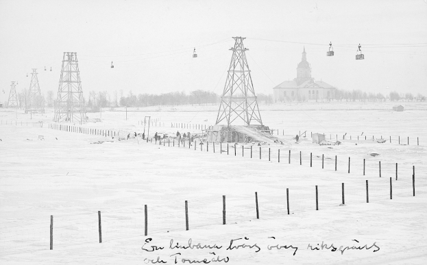 Ein Schwarz-Weiß-Foto einer Skiliftanlage in einer verschneiten Landschaft, umgeben von Pfählen, Seilen, Bäumen und einem Gebäude im Hintergrund, mit Text unten.
