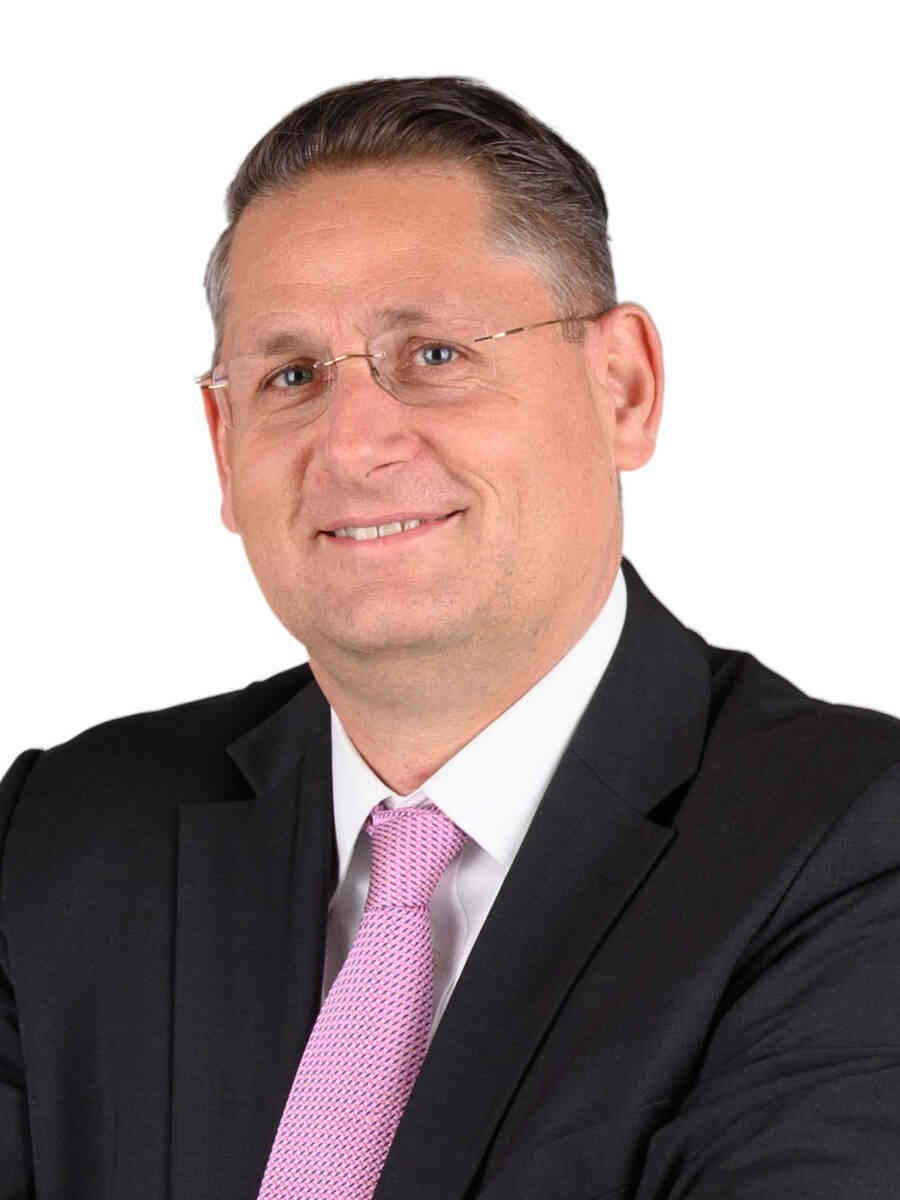Ein lachender Mann im Anzug, mit Krawatte und Brille vor einem weißen Hintergrund, wahrscheinlich ein Finanzberater.