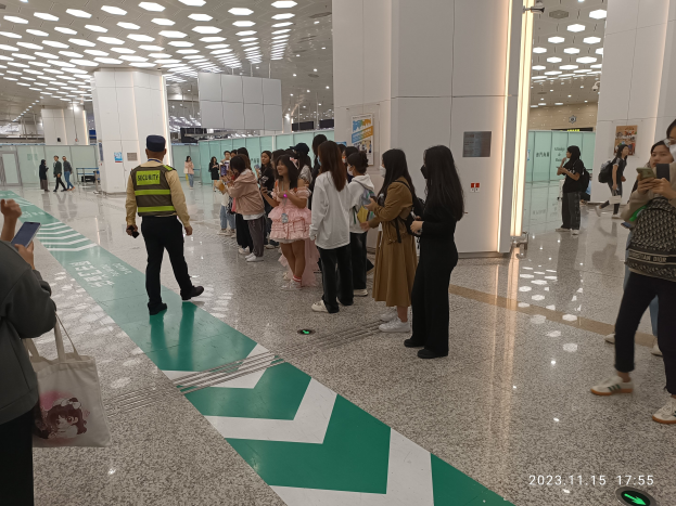 Eine Gruppe von Menschen, die in einer Reihe an einem Flughafen-Sicherheitscheck-in-Schalter stehen, einige halten Taschen und Mobiltelefone, mit Säulen und Deckenleuchten im Hintergrund.