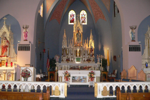 Innere einer Kirche mit hölzernen Kirchenbänken, Blumenarrangements, Kerzen auf Ständern, Statuen, Fahnen und Buntglasfenstern.