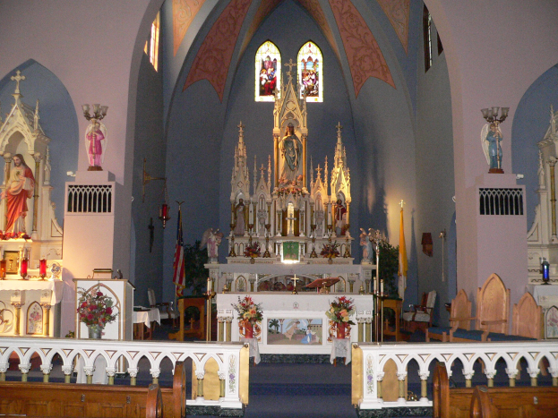 Innere einer Kirche mit hölzernen Kirchenbänken, Blumenarrangements, Kerzen auf Ständern, Statuen, Fahnen und Buntglasfenstern.