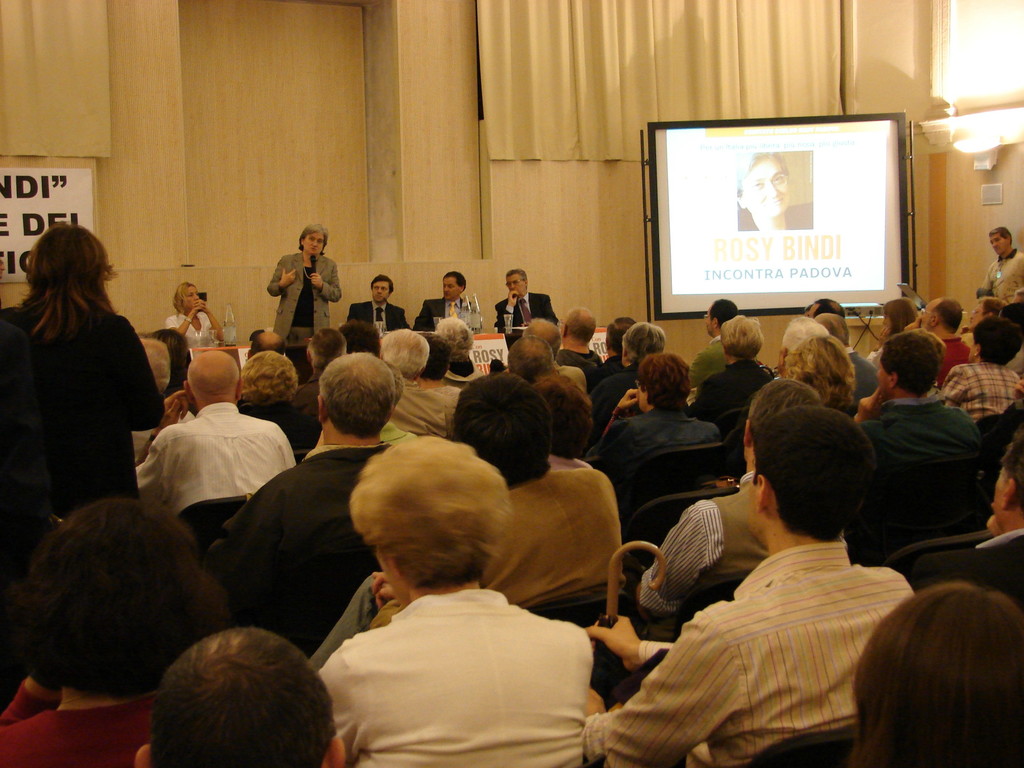 Eine Gruppe von Menschen, die auf Stühlen sitzen und einer Bühne mit einem Redner, der ein Mikrofon hält, sowie einem Bildschirm auf der rechten Seite und einem Vorhang oben zugewandt sind.