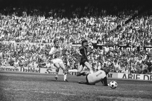 Schwarzes und weißes Foto von Fußballspielern im Einsatz auf einem Feld mit Zuschauern im Hintergrund und einer Bundesliga 1978-79 Spielankündigung.