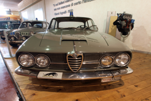 Ein 1970er Alfa Romeo Giulietta Sprint GT Junior in einem Museum ausgestellt, umgeben von Seilen und Pfosten, mit Text an der Wand und einer warm beleuchteten Decke.