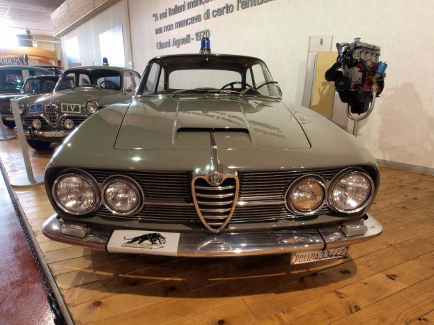 Ein 1970er Alfa Romeo Giulietta Sprint GT Junior in einem Museum ausgestellt, umgeben von Seilen und Pfosten, mit Text an der Wand und einer warm beleuchteten Decke.