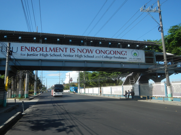 Ein Bus fährt unter einer Brücke mit einem "Jetzt ist Einschreibungszeit"-Schild, während Fußgänger darunter gehen und Stromleitungen entlang der Straße verlaufen, mit Bäumen und einem blauen Himmel im Hintergrund.