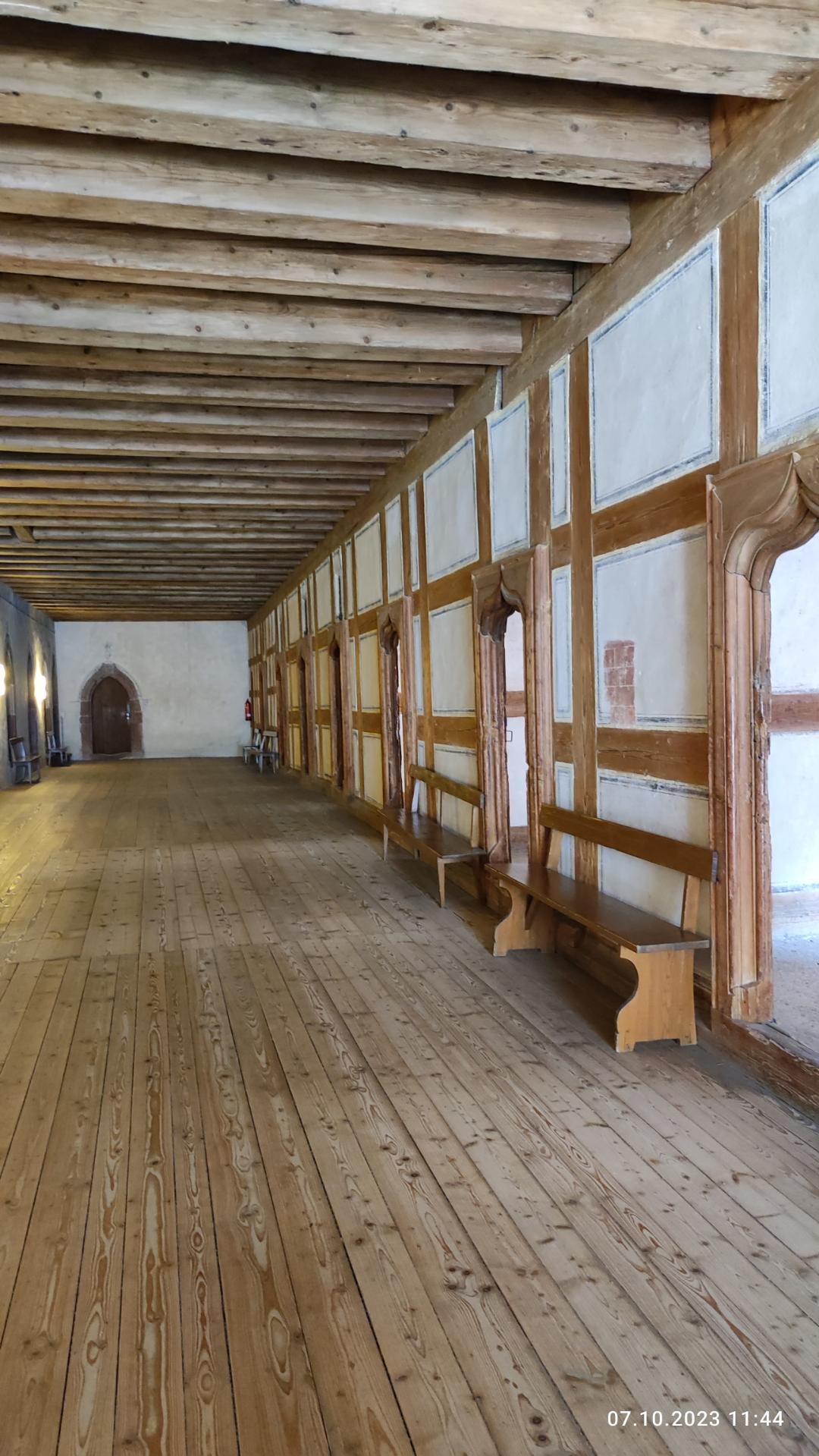 Ein geräumiger Raum mit Holzboden, weißen Wänden, Bänken, Fenstern, Lampen und einer hohen Decke, wahrscheinlich der Hauptsaal einer Burg.