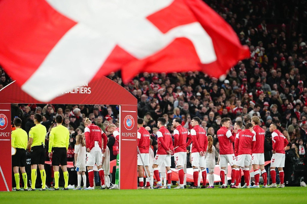 Eine Gruppe von Menschen auf einem Fußballfeld mit einer roten und weißen Flagge im Vordergrund, einem Bogen mit der Aufschrift "Bayern München vs Bayern Munich Betting Tips & Preview" im Hintergrund und einer großen Menschenmenge im Stadion.