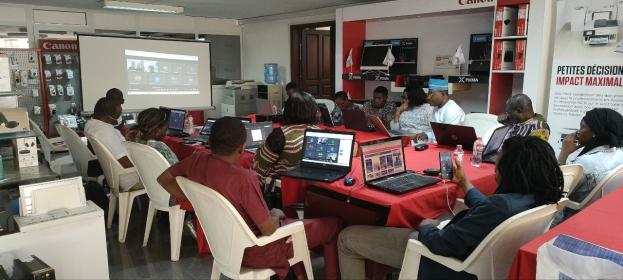 Gruppe von Menschen an einem Tisch mit Laptops und anderen Gegenständen während eines digitalen Marketing-Workshops, mit einem Banner, einem Display und Deckenleuchten im Raum.