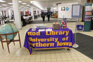 Eine Ausstellung mit einem Tisch, auf dem verschiedene Landesflaggen, eine Box, Papiere und ein Banner stehen, eine Kugel links daneben, ein Anschlagbrett rechts daneben mit ein paar Menschen in der Nähe und mehreren Tischen und Stühlen im Hintergrund.