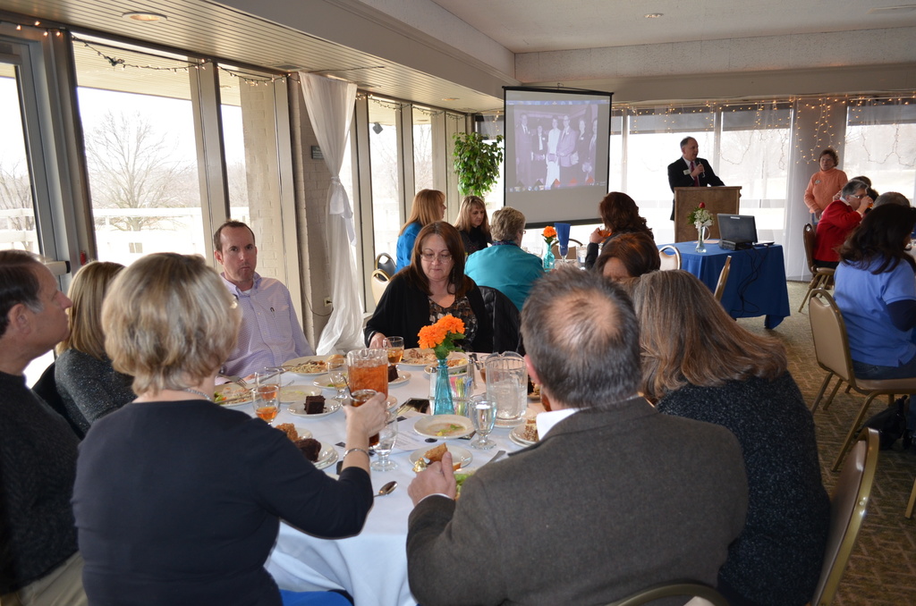 Eine Gruppe von Menschen an einem Tisch mit verschiedenen Speisen, eine Person steht und spricht, ein Projektor zeigt Inhalte, eine Pflanze daneben und ein Fenster mit Vorhängen, Himmel und Bäumen im Hintergrund.