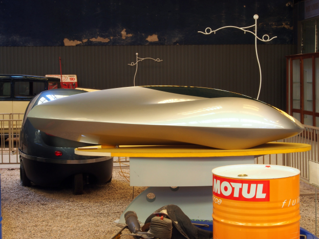 Kleines Hydrofoilboot auf einem Tisch neben einem Auto, mit zwei Trommeln auf dem Boden, einem Metallzaun und einer Glastür mit Text auf einer Tafel im Hintergrund.