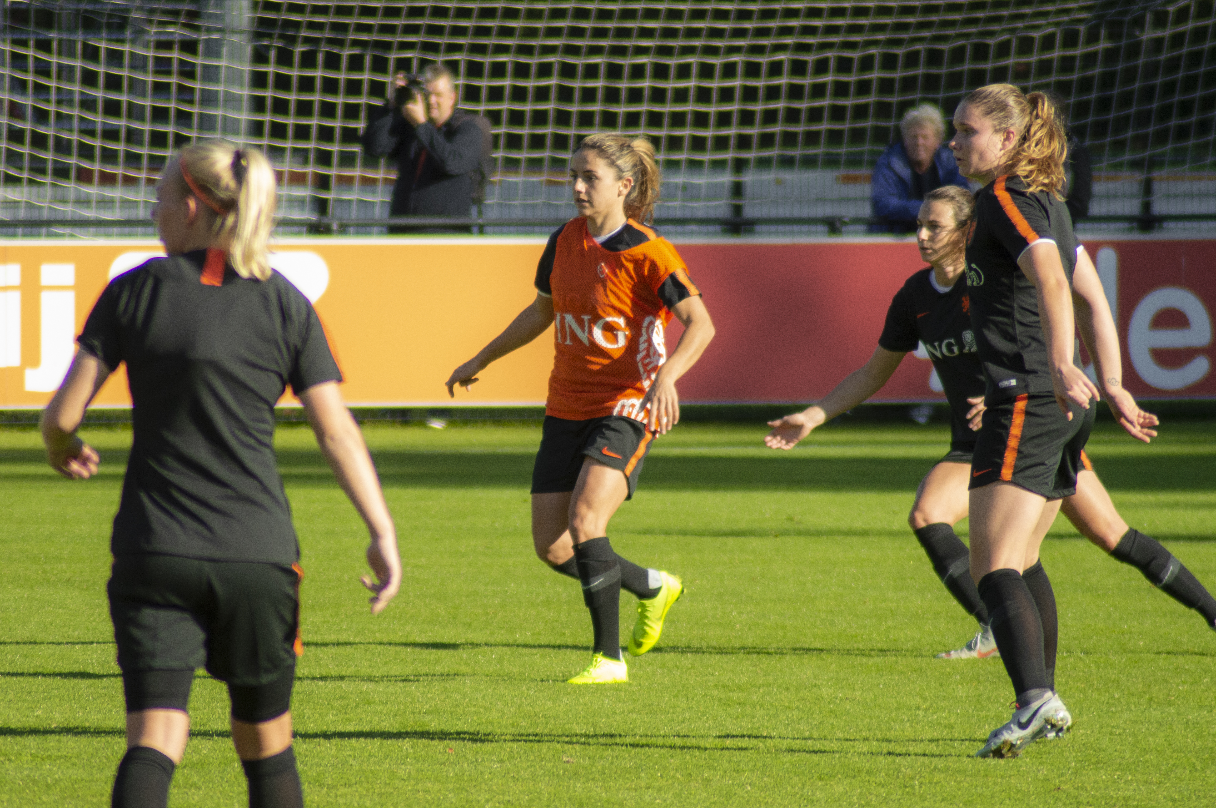 Eine Gruppe von Frauen, die auf einem Feld Fussball spielen, mit einem Netz im Hintergrund, Zuschauern auf B├Ąnken und einer Person mit einer Kamera, die die Aktion aufnimmt.