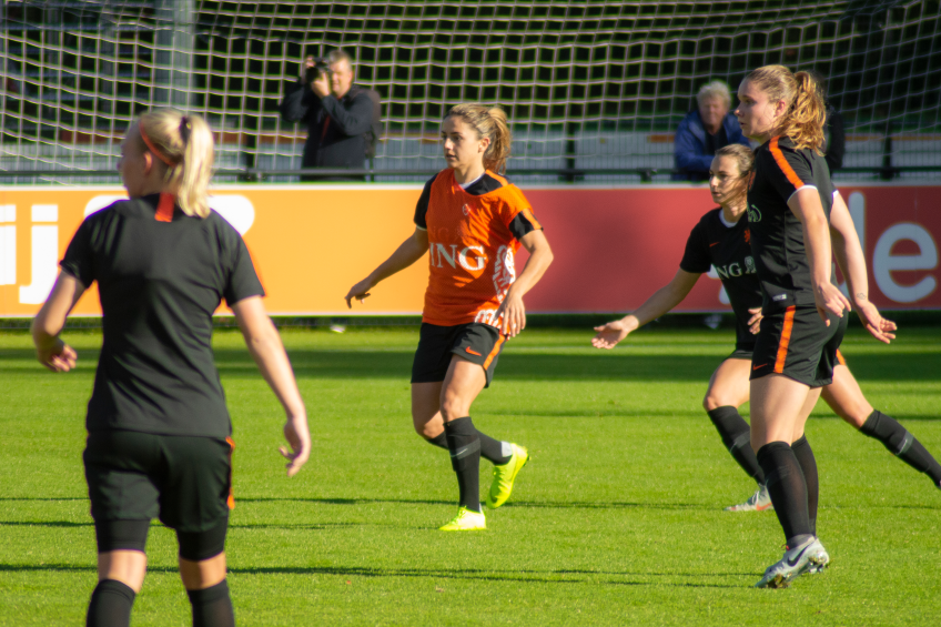 Eine Gruppe von Frauen, die auf einem Feld Fussball spielen, mit einem Netz im Hintergrund, Zuschauern auf B├Ąnken und einer Person mit einer Kamera, die die Aktion aufnimmt.
