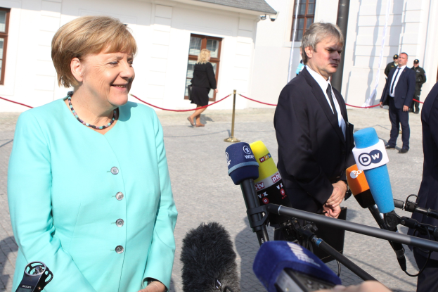Bundeskanzlerin Angela Merkel beim Sprechen mit Reportern vor einem Gebäude mit einer Gruppe von Menschen und Mikrofonen unter einem bewölkten Himmel.
