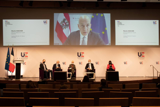 Eine Gruppe von Menschen, die auf einer Bühne in einem Halbkreis um ein zentrales Podium mit Mikrofon sitzen, flankiert von Fahnen, mit einem Bildschirm im Hintergrund, der Text und Bilder anzeigt, während einer Pressekonferenz der Europäischen Union.